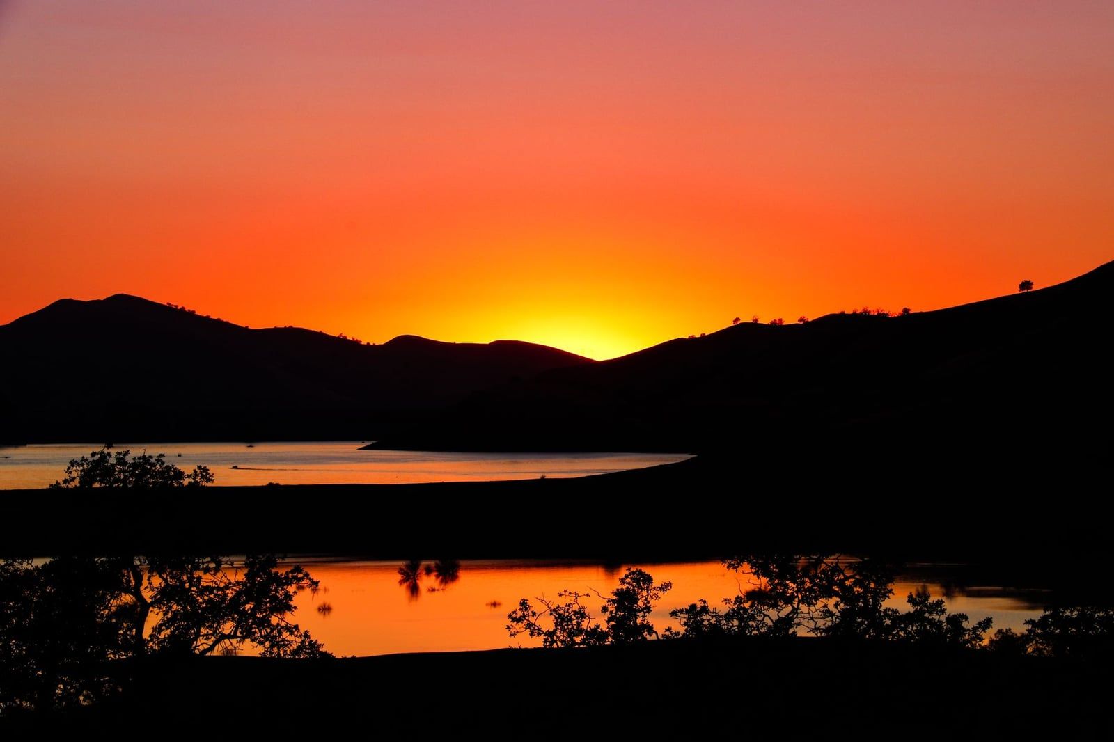 A brilliant orange sunset reflects across Lake Kaweah amid dark rolling hills in Three Rivers, California, near Sequoia National Park.