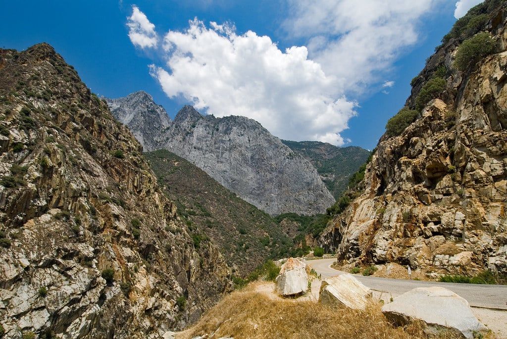 Eine asphaltierte Kurve des Kings Canyon Scenic Byway schlängelt sich entlang hoher Granit-Canyonwände unter blauem Himmel mit weißen Wolken.