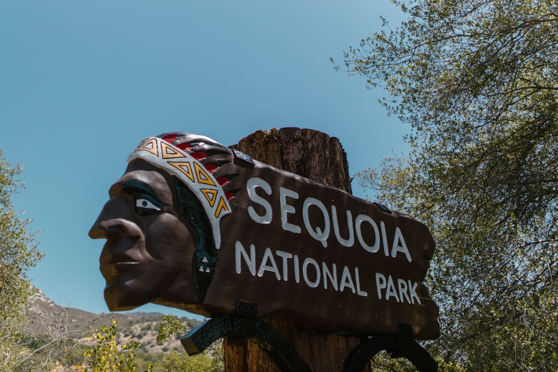 A carved wooden entrance sign featuring a Native American profile welcomes visitors to Sequoia National Park under a clear blue sky.