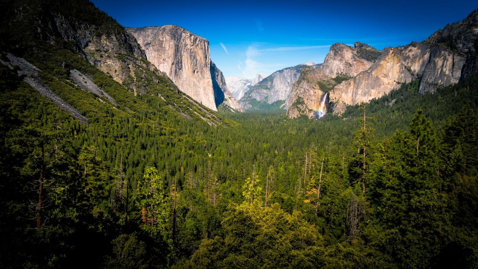 Panoramic vista of Yosemite Valley from Tunnel View, featuring El Capitan, Half Dome, and Bridalveil Fall with a rainbow.
