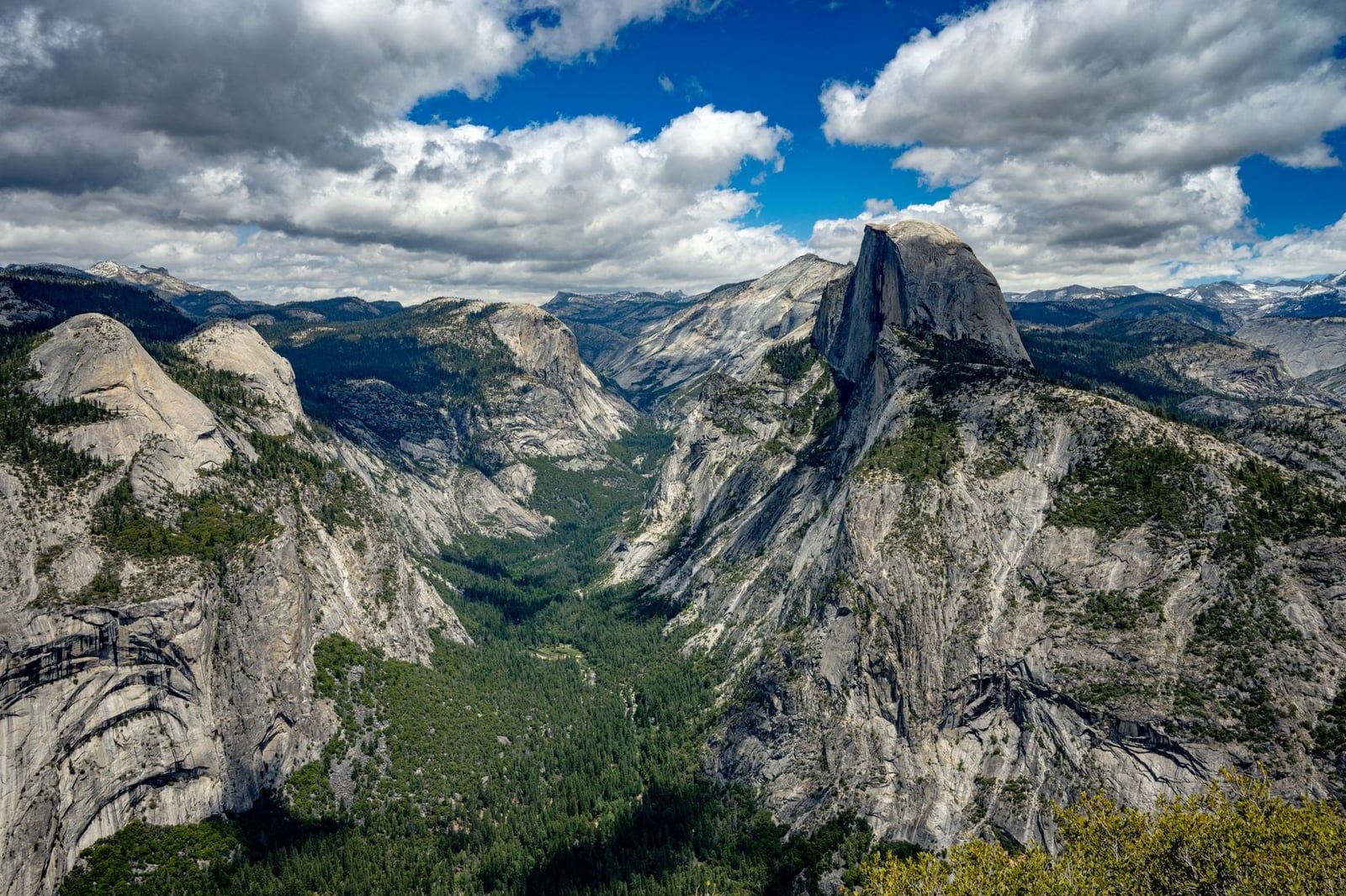 Panoramic view of Half Dome rising above the deep granite valleys and pine forests of Yosemite National Park under a partly cloudy sky.